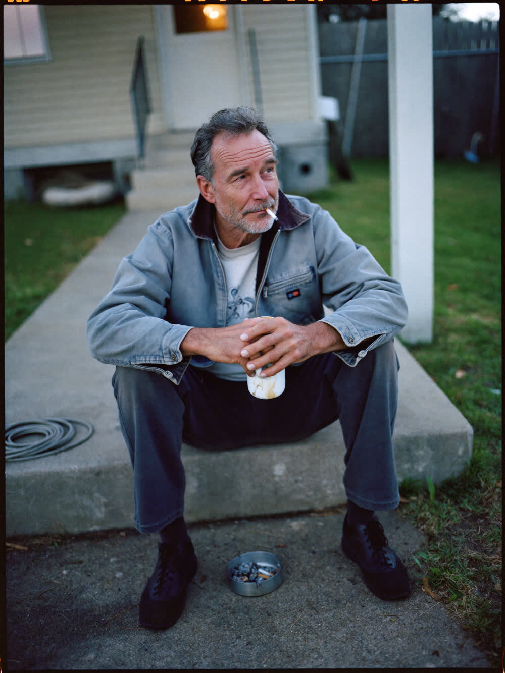 A man in his early sixties sitting on his back step at dawn with a coffee mug, an ashtray on the step beside him
